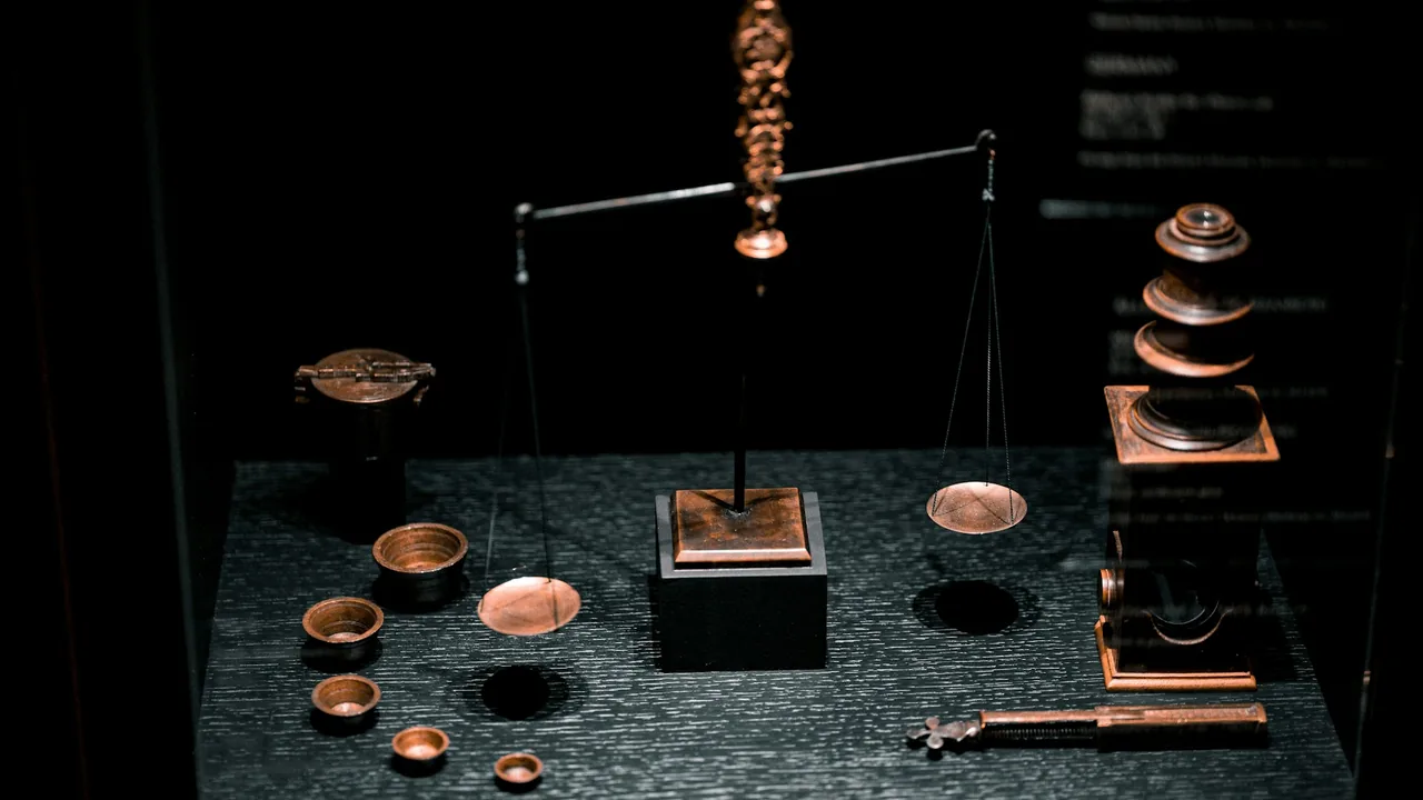 Koyu bir zeminde bakır kefeli antika bir terazi ve çevresine dizilmiş ağırlıklar